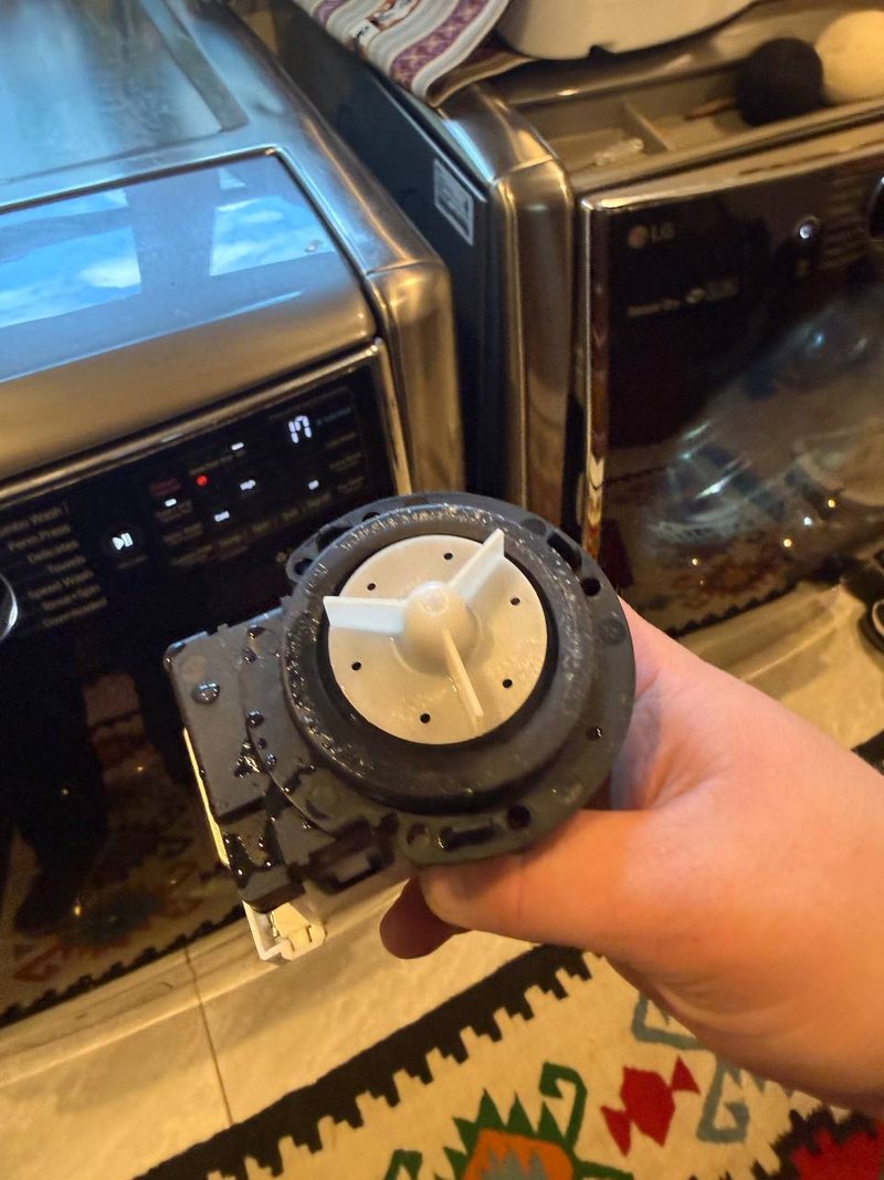 Drain pump removed from an LG front-load washer, held up in front of the washer and dryer pair
